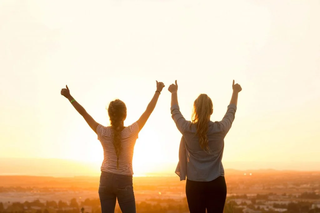 deux femmes de dos devant un coucher de soleil levant les pouces en l'air